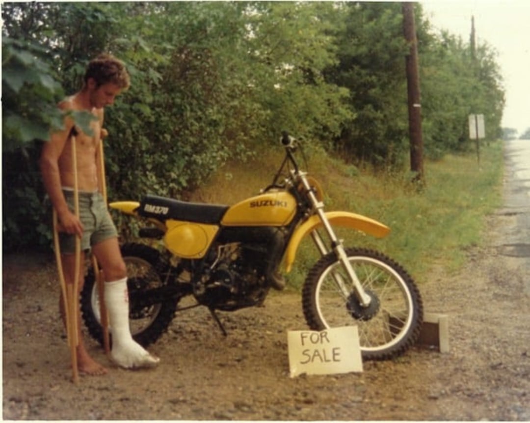 A man with a cast on his leg and crutches stands next to a yellow Suzuki dirt bike with a "FOR SALE" sign, beside a rural road with trees and grass.