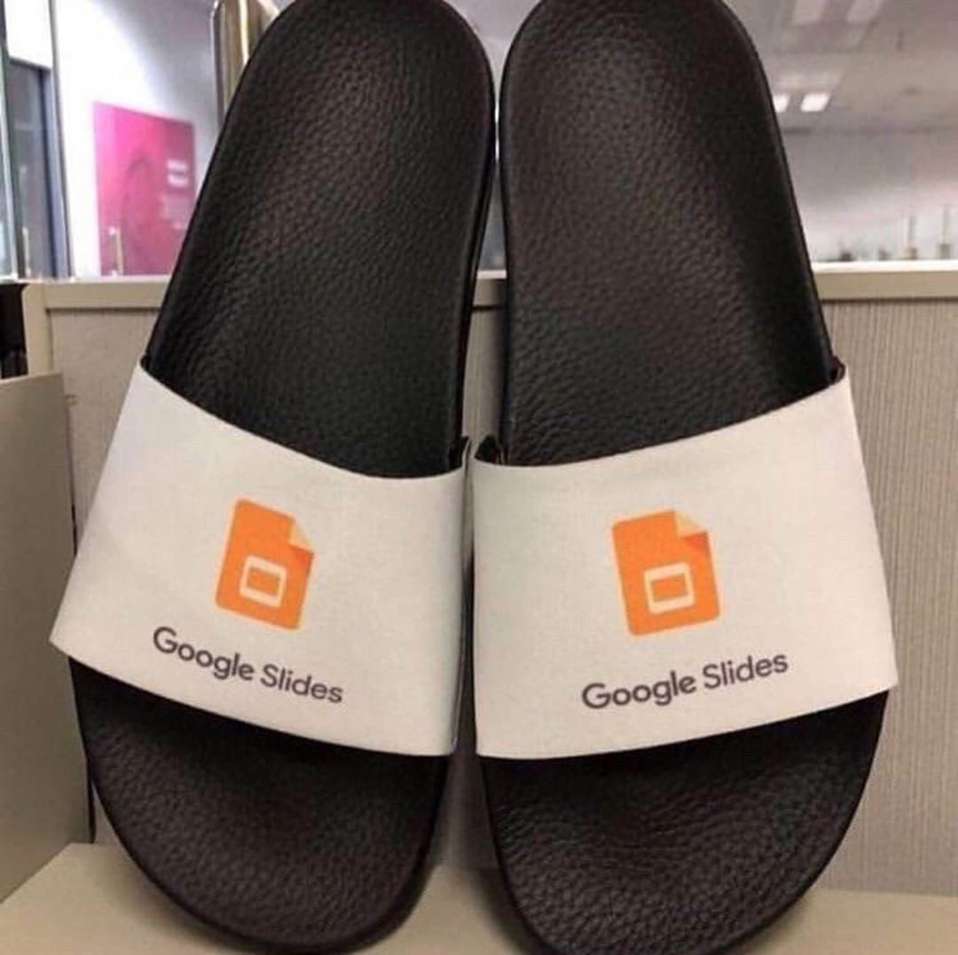 A pair of black slide sandals with white straps, each featuring the Google Slides logo and the text "Google Slides" printed on the strap.