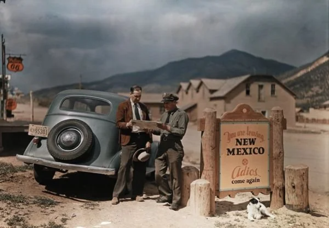Two men stand by a vintage car, looking at a map near a sign that reads, “You are leaving New Mexico. Adios, come again.” Mountains and buildings are in the background; a small dog sits nearby.