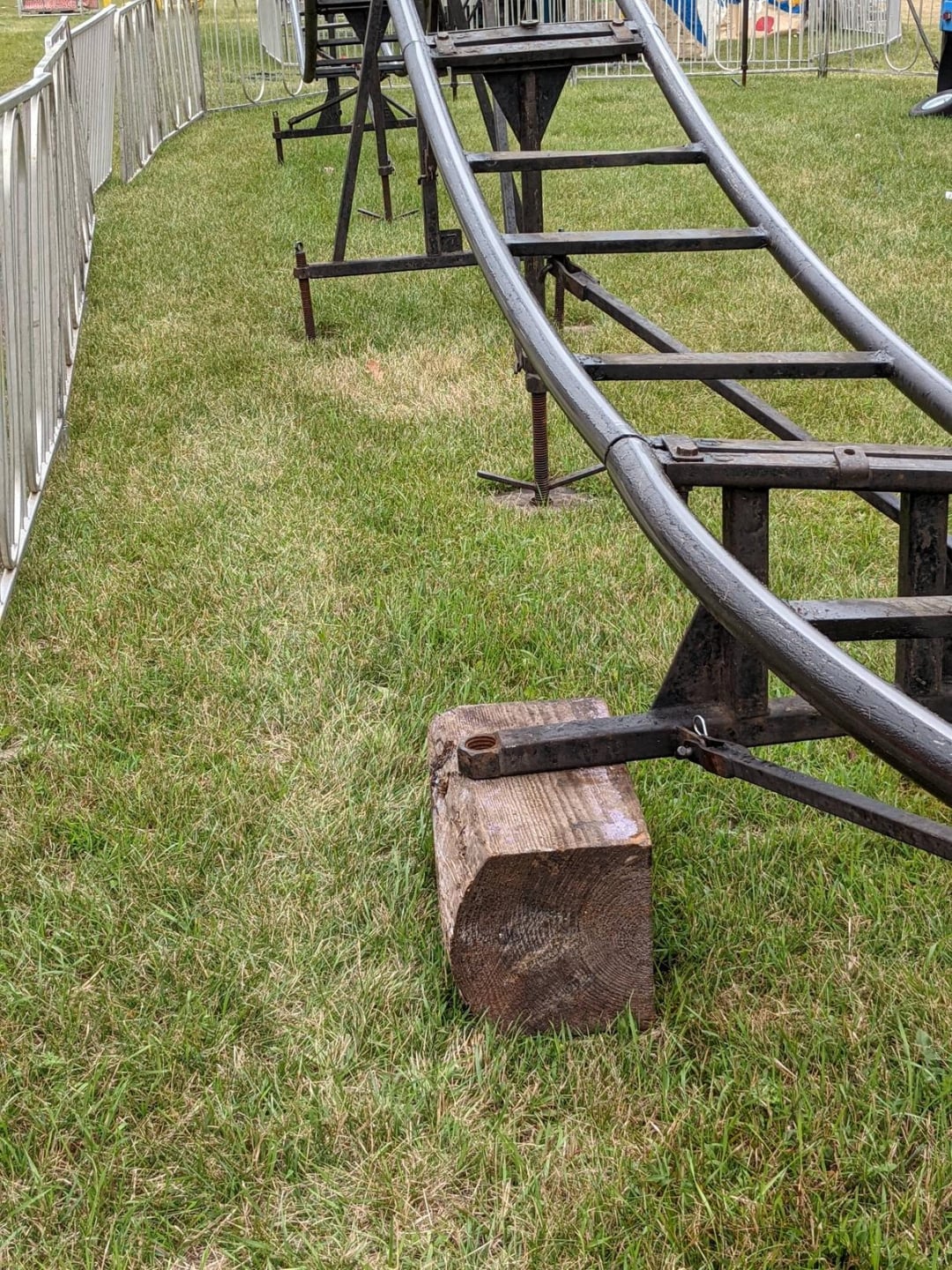 A close-up of a metal roller coaster track on grass, supported by a wooden block and metal frame, with white safety railings on both sides.