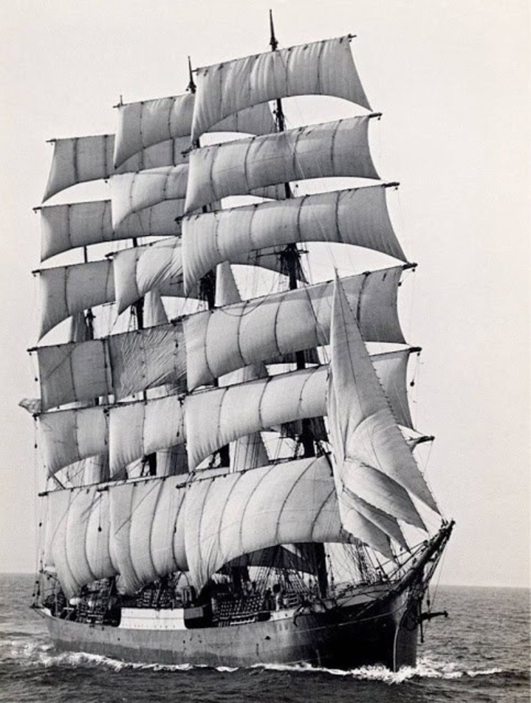 A large, historic sailing ship with multiple masts and numerous sails unfurled, moving forward on the open sea. The ship appears to be a classic tall ship or clipper, photographed in black and white.