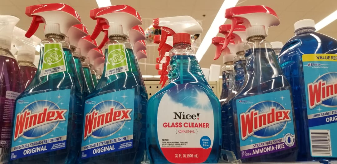 Bottles of Windex and Nice! glass cleaner are displayed on a store shelf. The Windex bottles have blue liquid and red-and-white spray tops, while the Nice! cleaner has a red spray top and a white label.