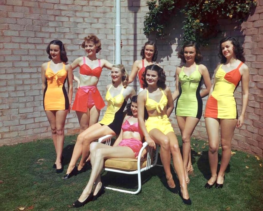 Seven women in colorful vintage swimsuits pose outdoors by a brick wall, with two seated on a chair and five standing behind them on grass. Bright sunlight highlights their smiling faces and vibrant outfits.