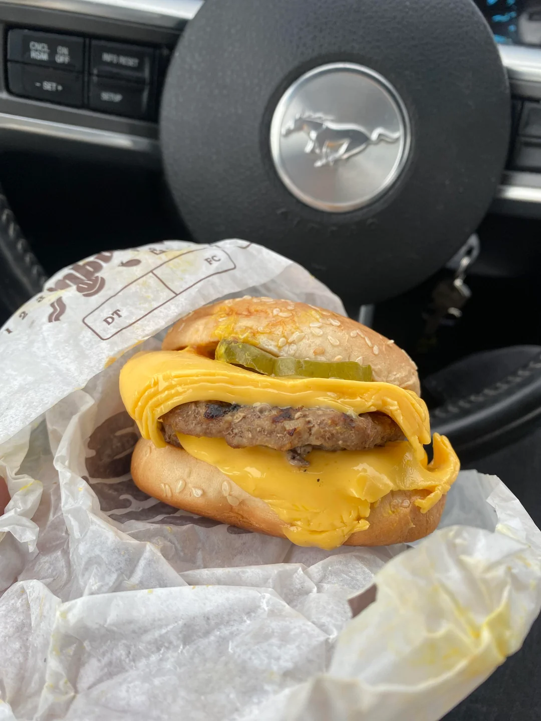 A cheeseburger with melted cheese, pickles, and a beef patty is wrapped in paper and held inside a car, with a Mustang steering wheel visible in the background.