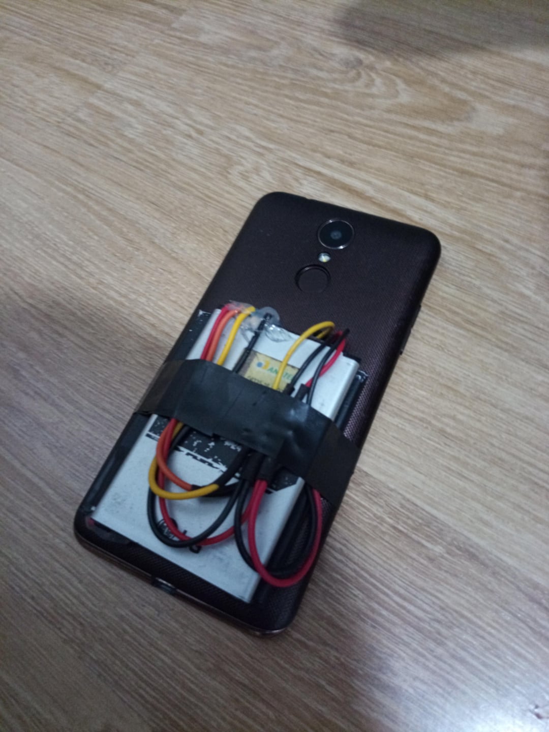 A smartphone lies face down on a wooden surface with electronic components and wires taped to its back using black tape.