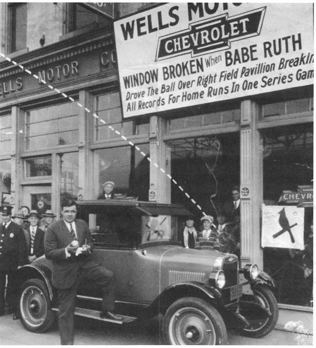 Black-and-white photo of Babe Ruth posing with a bat and a car under a sign at Wells Motor Co., noting a broken window caused by Ruth hitting a home run. Several men in hats stand nearby, and the building is labeled Chevrolet.