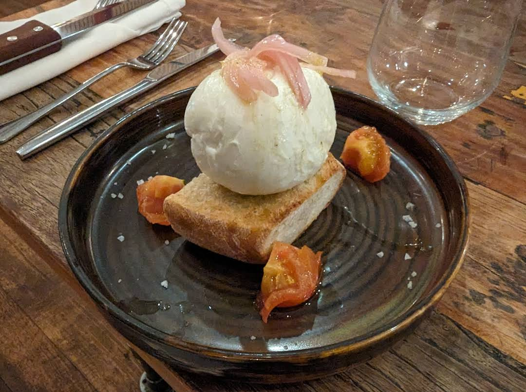 A round ball of burrata cheese sits on a piece of toasted bread, garnished with pickled onions and surrounded by quartered cherry tomatoes on a dark ceramic plate. Silverware and a glass are in the background.