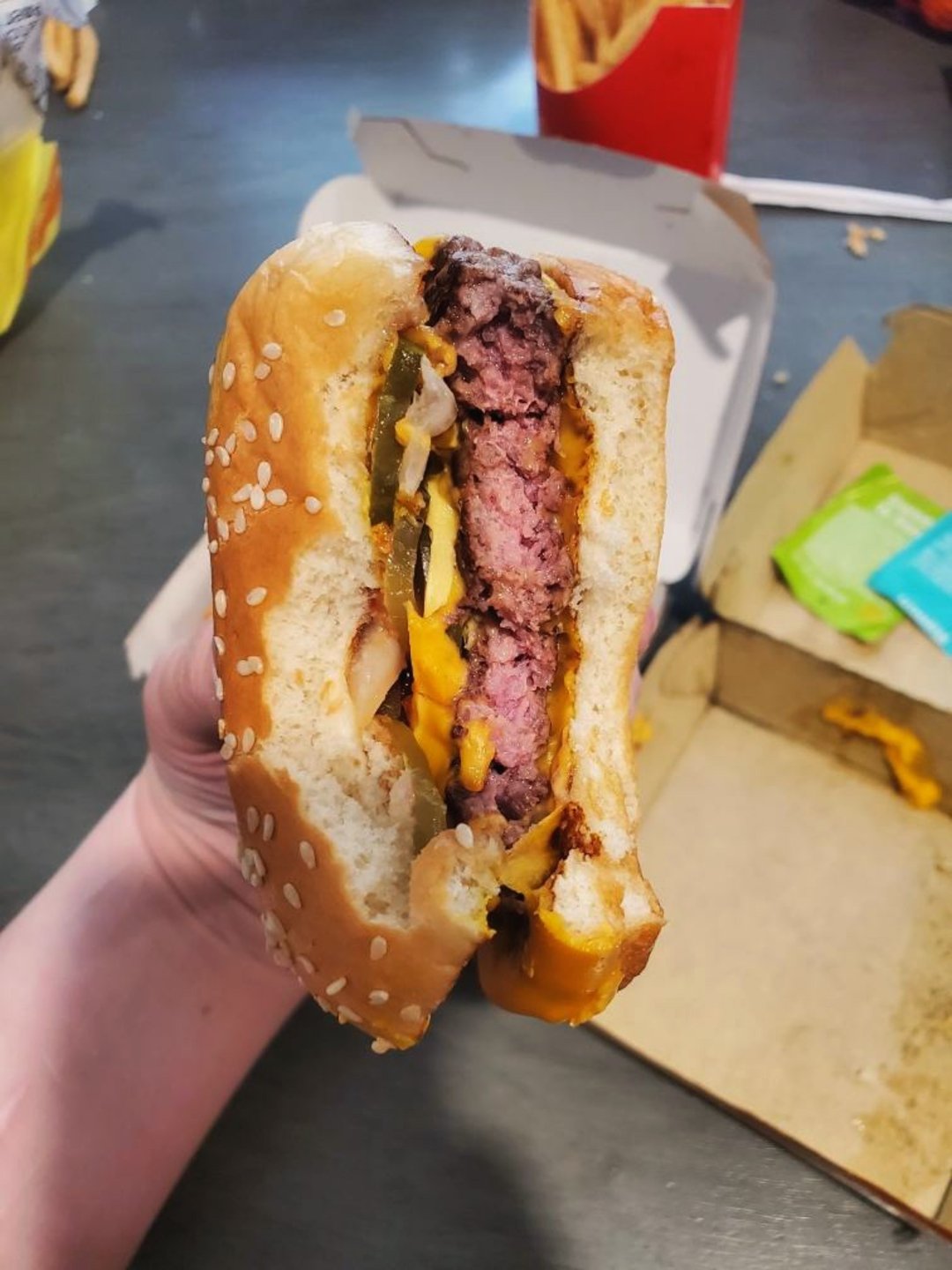 A hand holds a bitten cheeseburger with a sesame seed bun, showing a thick beef patty, melted cheese, onions, and pickles. In the background are fries in a red carton and an open food box.