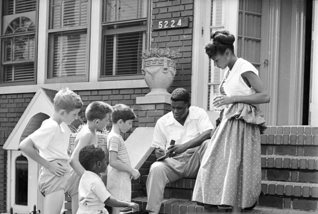 A man sits on brick steps outside a house numbered 5224, showing something to four children, while a woman stands nearby holding a drink. The group appears engaged and relaxed in front of a brick building.