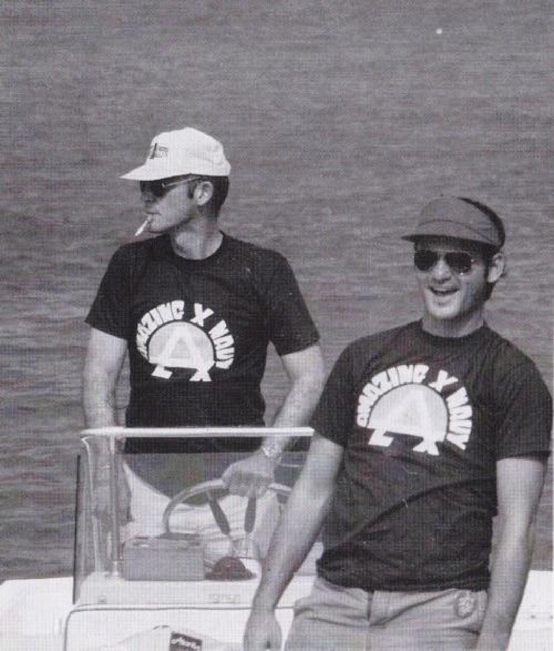 Two men stand on a boat wearing matching "Crazing Y Idak" t-shirts. One is steering with a cigarette in his mouth; the other is smiling broadly. Both wear sunglasses and caps. The water is calm in the background.