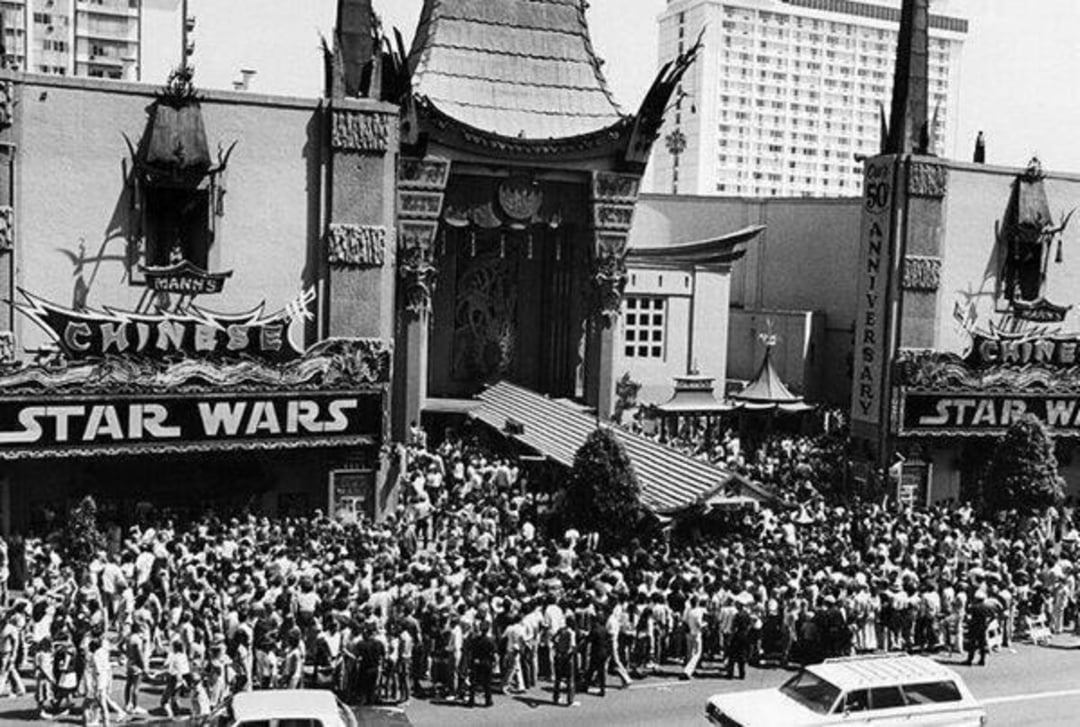 A large crowd gathers outside Grauman’s Chinese Theatre in Los Angeles beneath a marquee advertising "Star Wars," during its 1977 premiere; cars are parked in front and a tall building stands in the background.