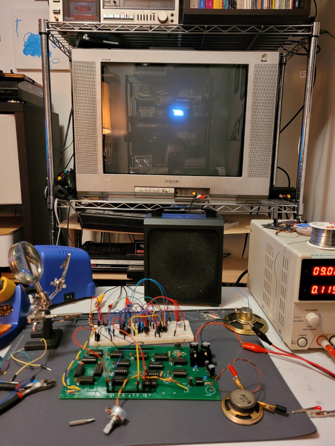 A home electronics workspace with a breadboard circuit, tools, wires, and multimeter on a desk in front of an old CRT television displaying a small blue square. Shelves with more equipment are in the background.
