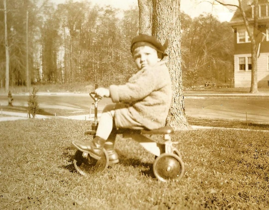 A young child wearing a coat and hat rides a tricycle on grass near a large tree, with a house and road visible in the background. The photo is in sepia tone, giving it a vintage feel.