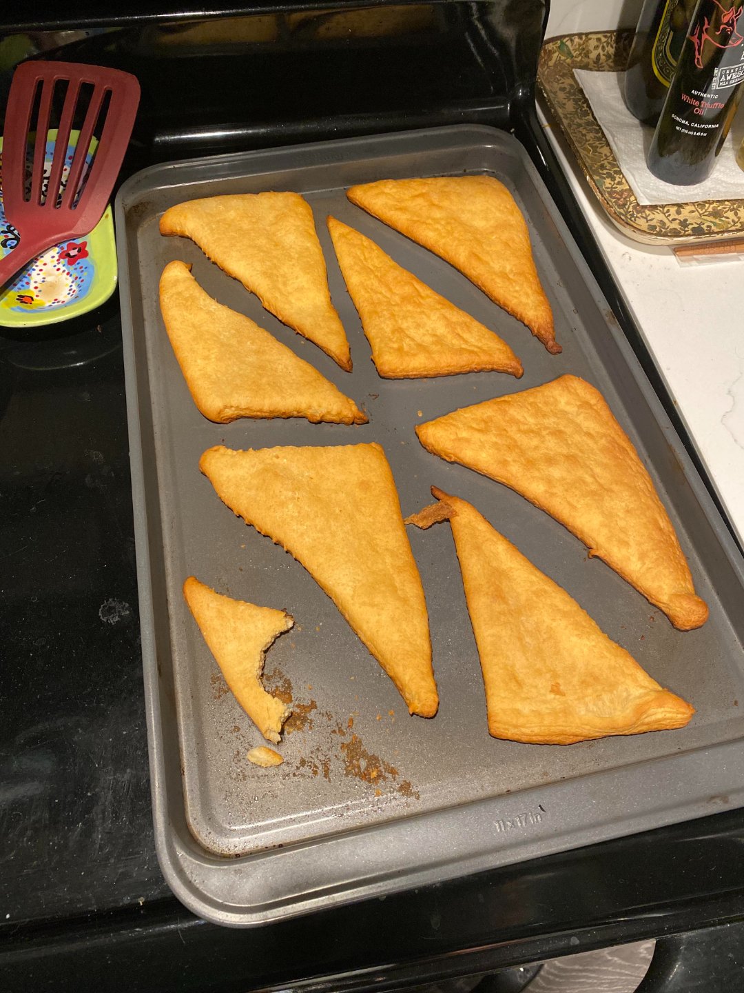 Baked golden-brown crescent roll dough pieces are arranged on a baking sheet on a stovetop. One piece has a bite taken out of it. Kitchen utensils and other items are visible nearby.