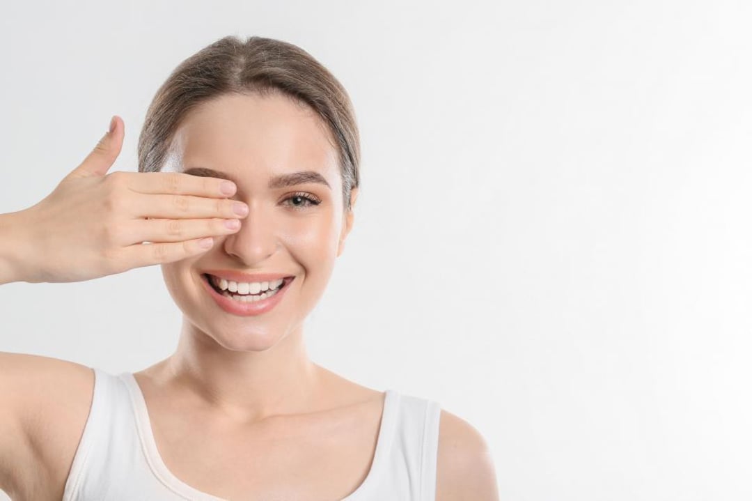 A smiling young woman with brown hair in a white tank top covers one eye with her hand, standing against a plain light background.