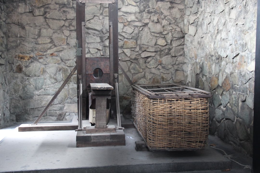 A wooden guillotine stands next to a large, rectangular wicker basket in a stone-walled room, creating a somber, historical atmosphere.