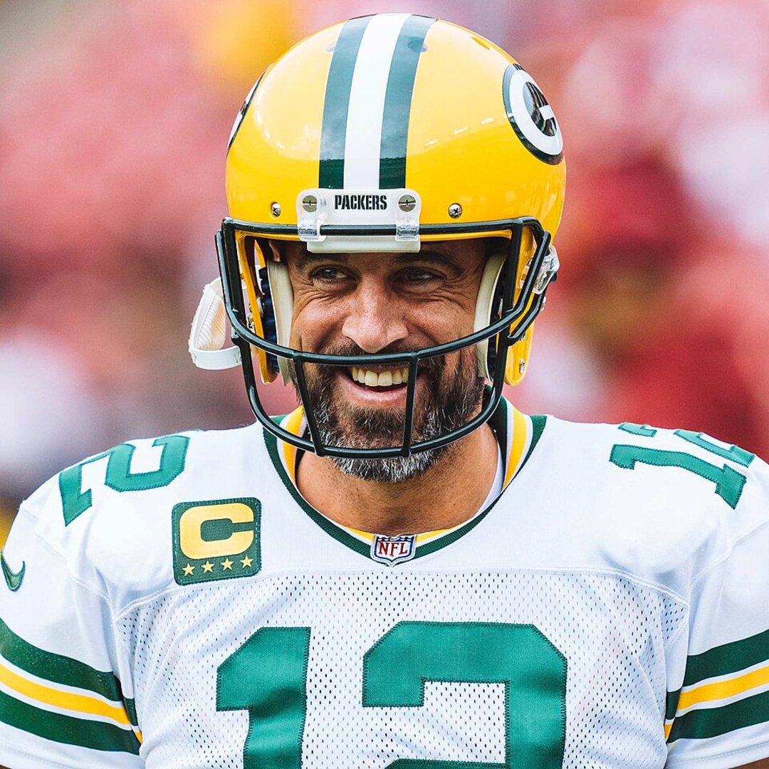 A football player wearing a yellow helmet and a Green Bay Packers uniform with the number 12 smiles on the field.