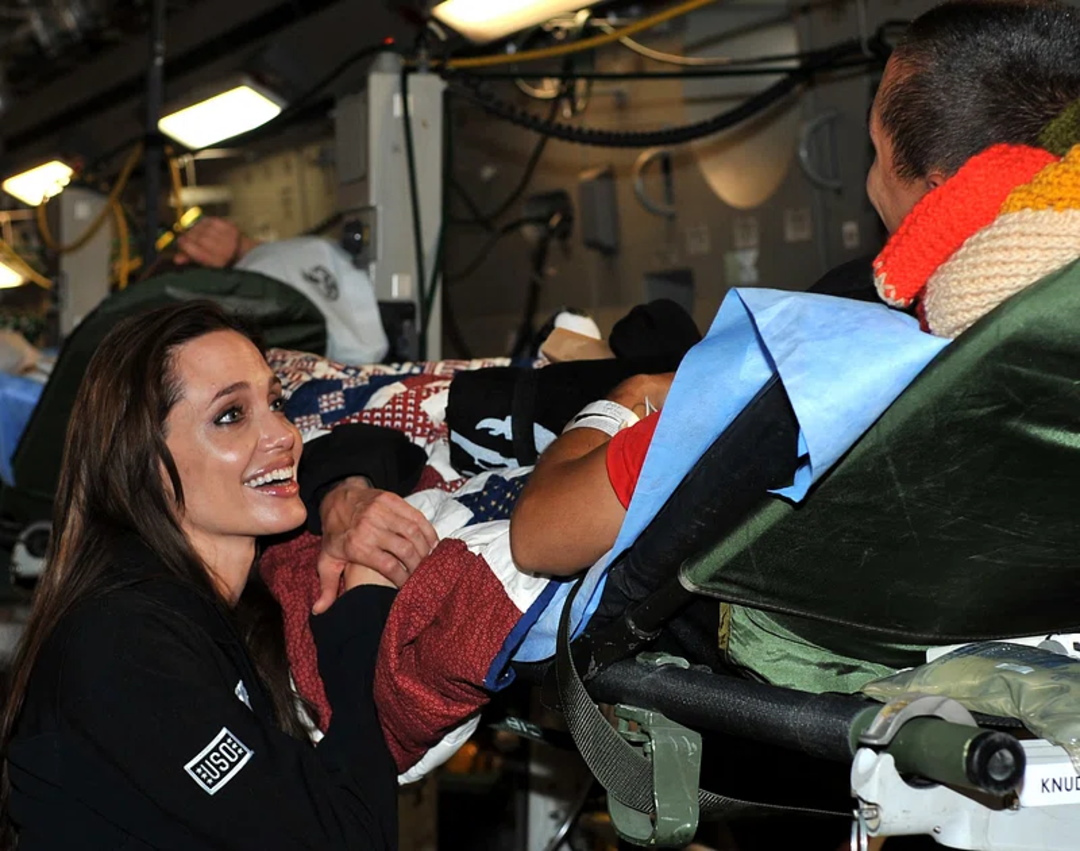 A woman smiling and holding hands with a patient lying on a stretcher covered by a patriotic blanket, inside what appears to be a medical or military transport setting.