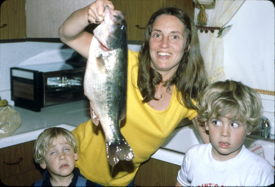 A smiling woman in a yellow shirt holds up a large fish in a kitchen, while two young children stand beside her with surprised and amused expressions.
