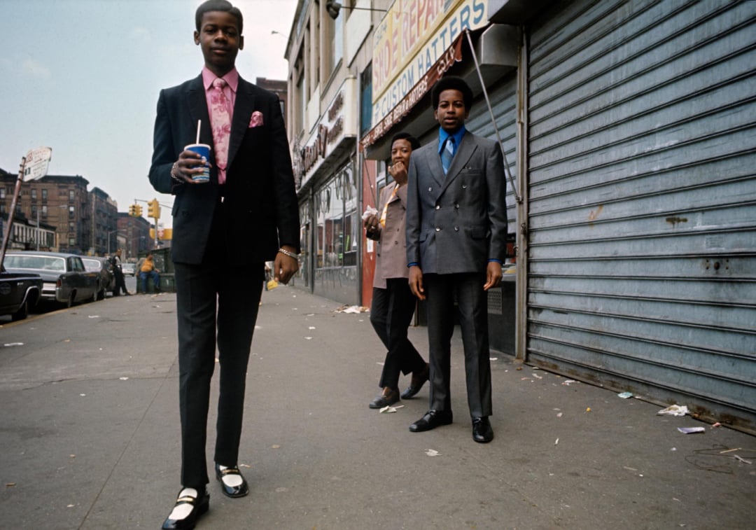 Three young men in stylish suits stand on a city sidewalk; one walks towards the camera holding a drink, while the other two stand near a closed storefront with metal shutters. Urban buildings and cars line the street.
