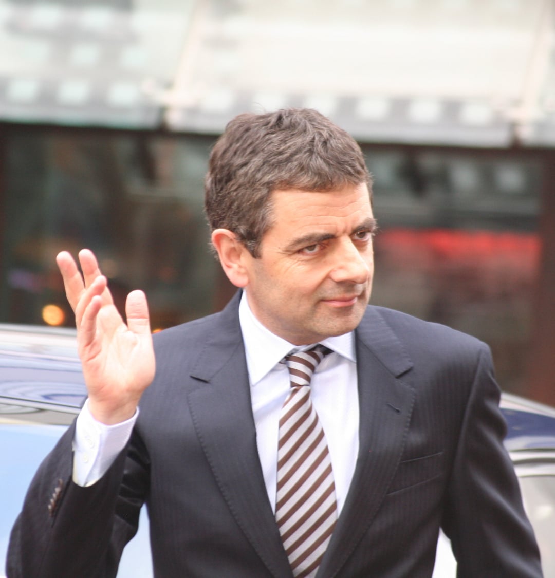 A man in a suit and striped tie stands outdoors, raising his hand in a wave. He has short brown hair and is looking slightly to the side with a neutral expression. A blurred background includes a car and glass structure.