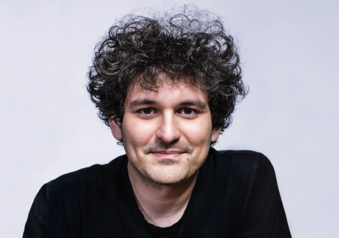 A person with curly dark hair and a slight smile is wearing a black shirt against a plain light-colored background.