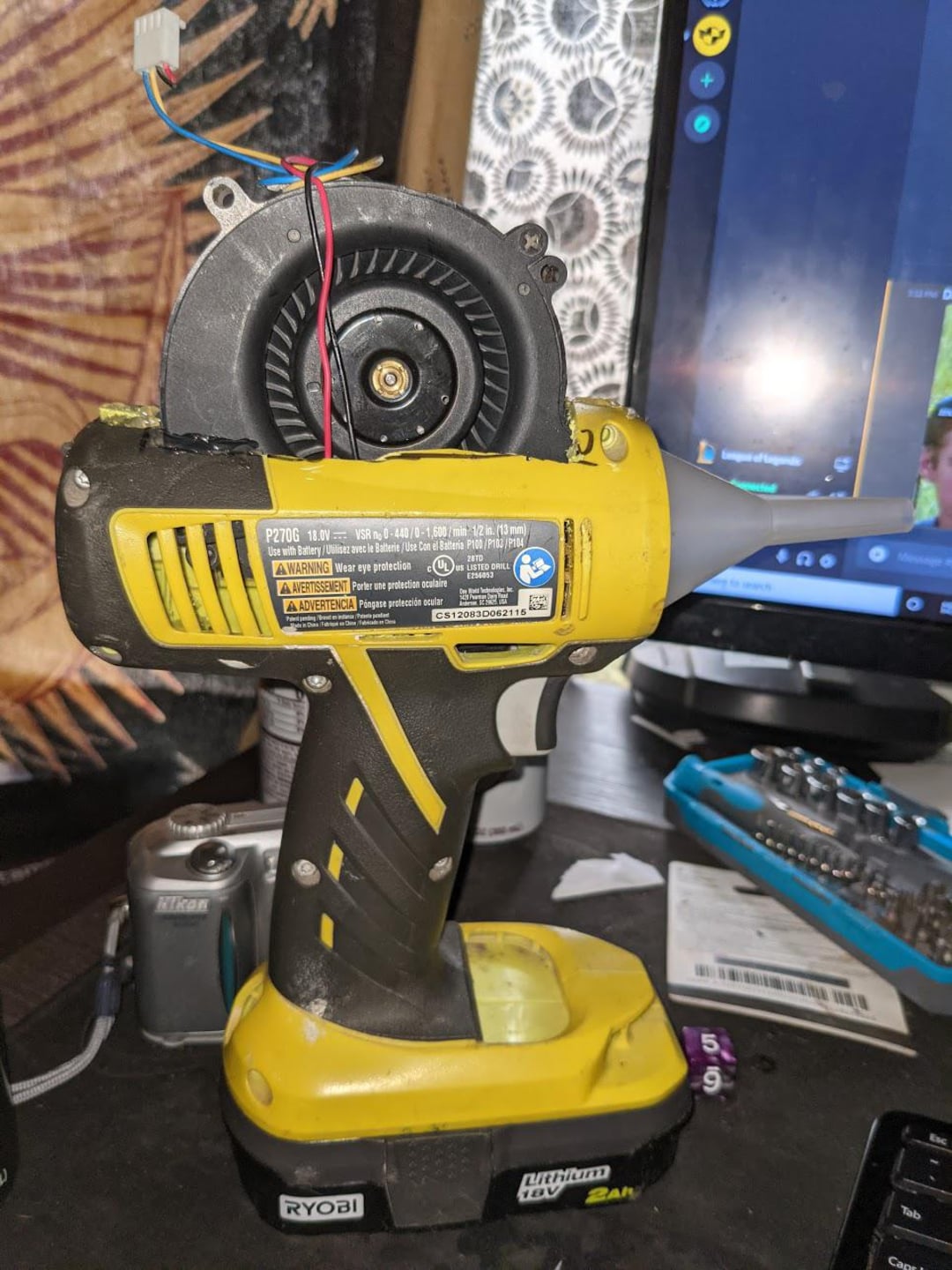 A handheld Ryobi power tool modified with a fan and nozzle attachment on top, sitting on a cluttered desk near a computer monitor, camera, and various electronic items.