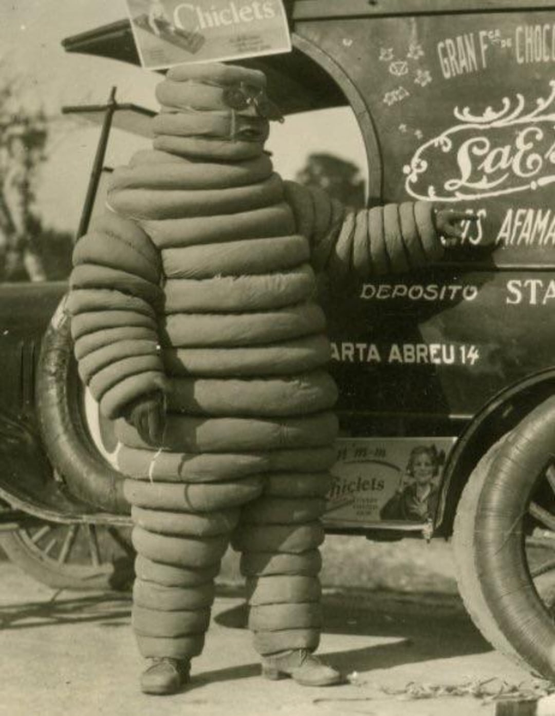 A person dressed in a vintage Michelin Man costume, made of stacked, rounded segments, stands next to an old car covered with advertising text and posters.