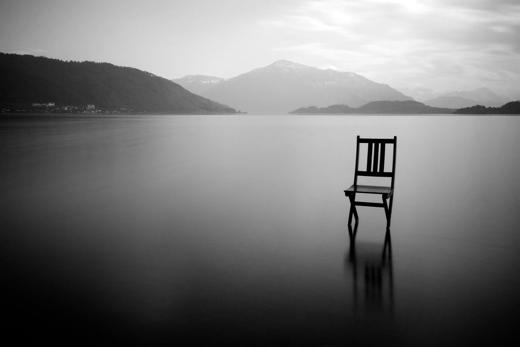 A solitary wooden chair stands in shallow water, facing distant mountains under a cloudy sky. The scene is calm and monochromatic, evoking a sense of solitude and tranquility.