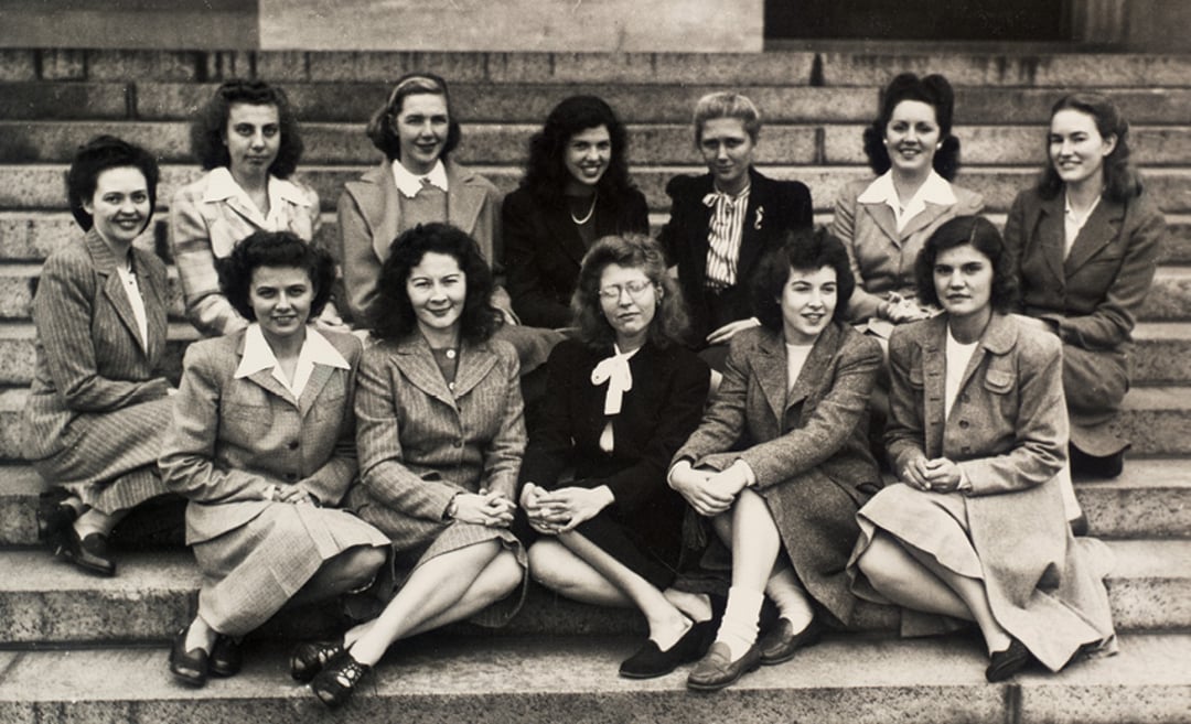 Black and white photo of eleven women in vintage attire, sitting and kneeling on outdoor steps, posing together and smiling for the camera.