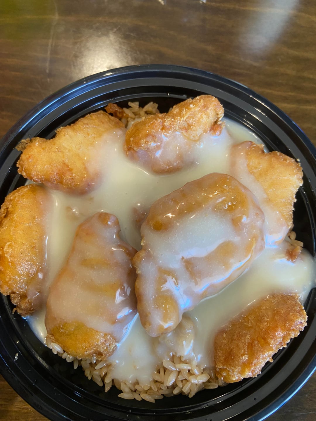A black bowl filled with fried chicken pieces on a bed of rice, covered in a thick, creamy white sauce, placed on a wooden table.