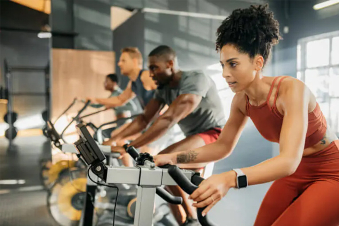 A group of people are focused and exercising on stationary bikes in a gym, with a woman in the foreground wearing red athletic wear and a smartwatch. Sunlight streams in through windows.