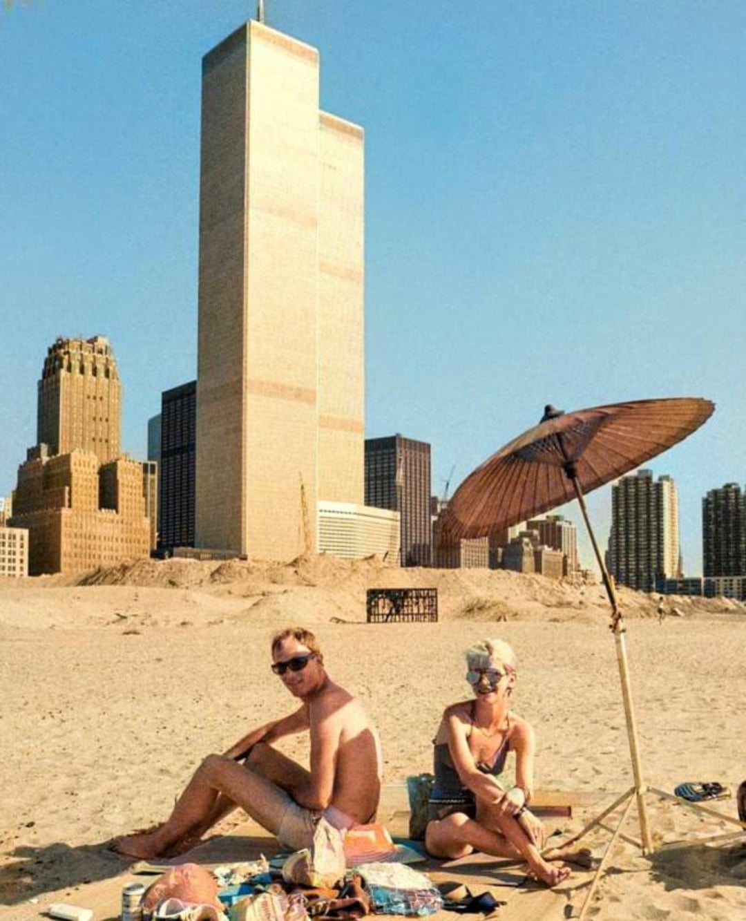 Two people sit on a sandy beach under a sun umbrella, with the Twin Towers of the World Trade Center and other New York City buildings visible in the background under a clear blue sky.