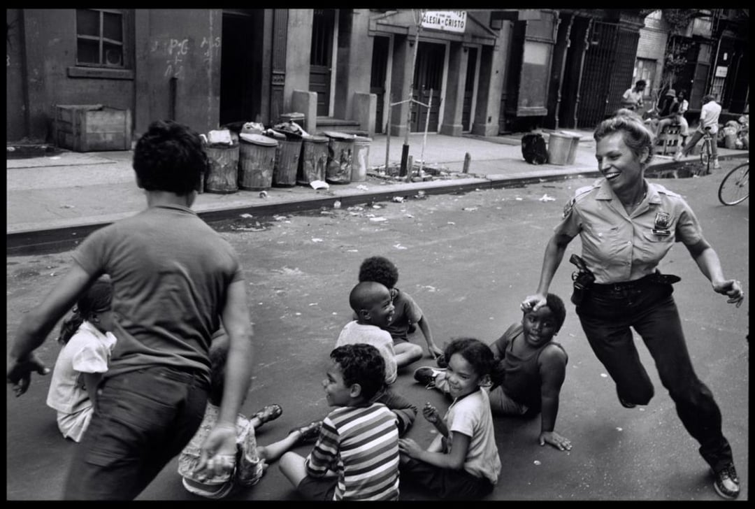 A police officer playfully chases laughing children who are sitting on a city street. The scene is energetic, with other kids and urban buildings, trash cans, and people visible in the background.