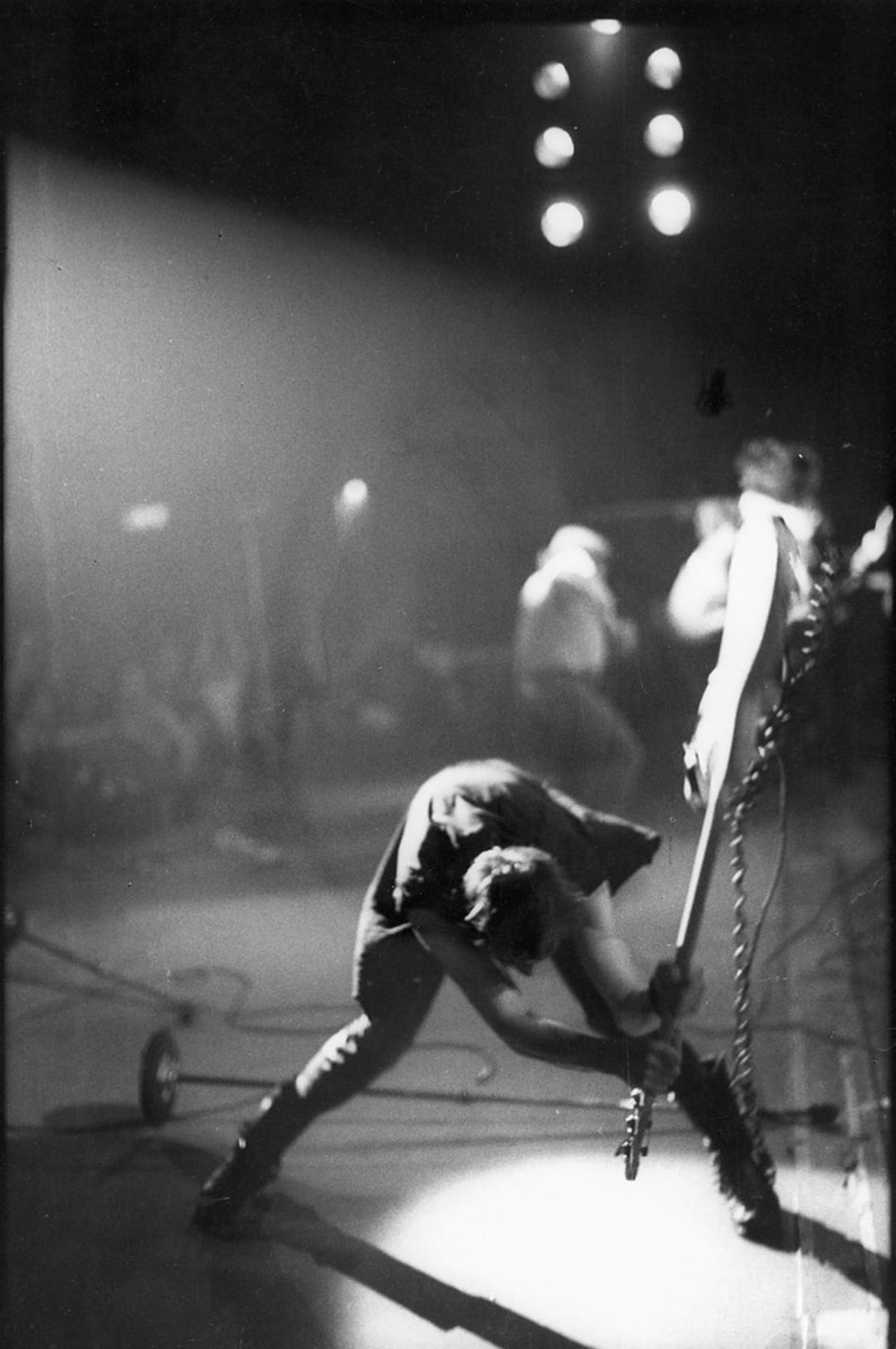 A black-and-white photo of a musician mid-action, swinging a guitar downward on stage. Bright spotlights shine behind, with other band members and musical equipment visible in the background.