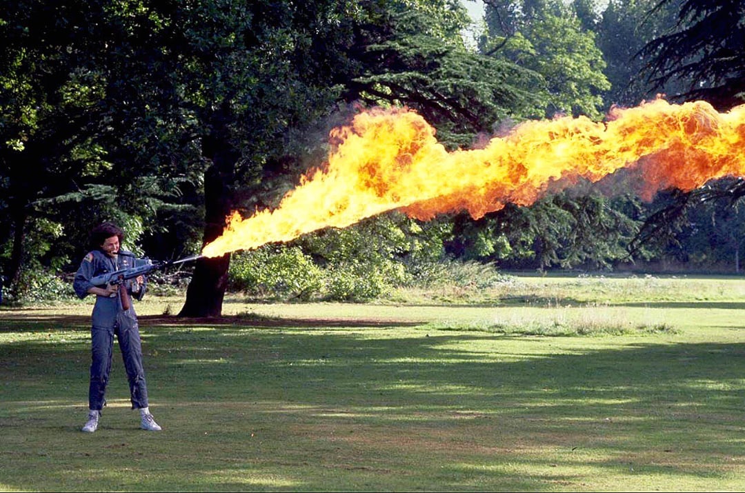 A person standing on grass outdoors uses a flamethrower, shooting a large stream of fire forward. Lush green trees and bushes fill the background.