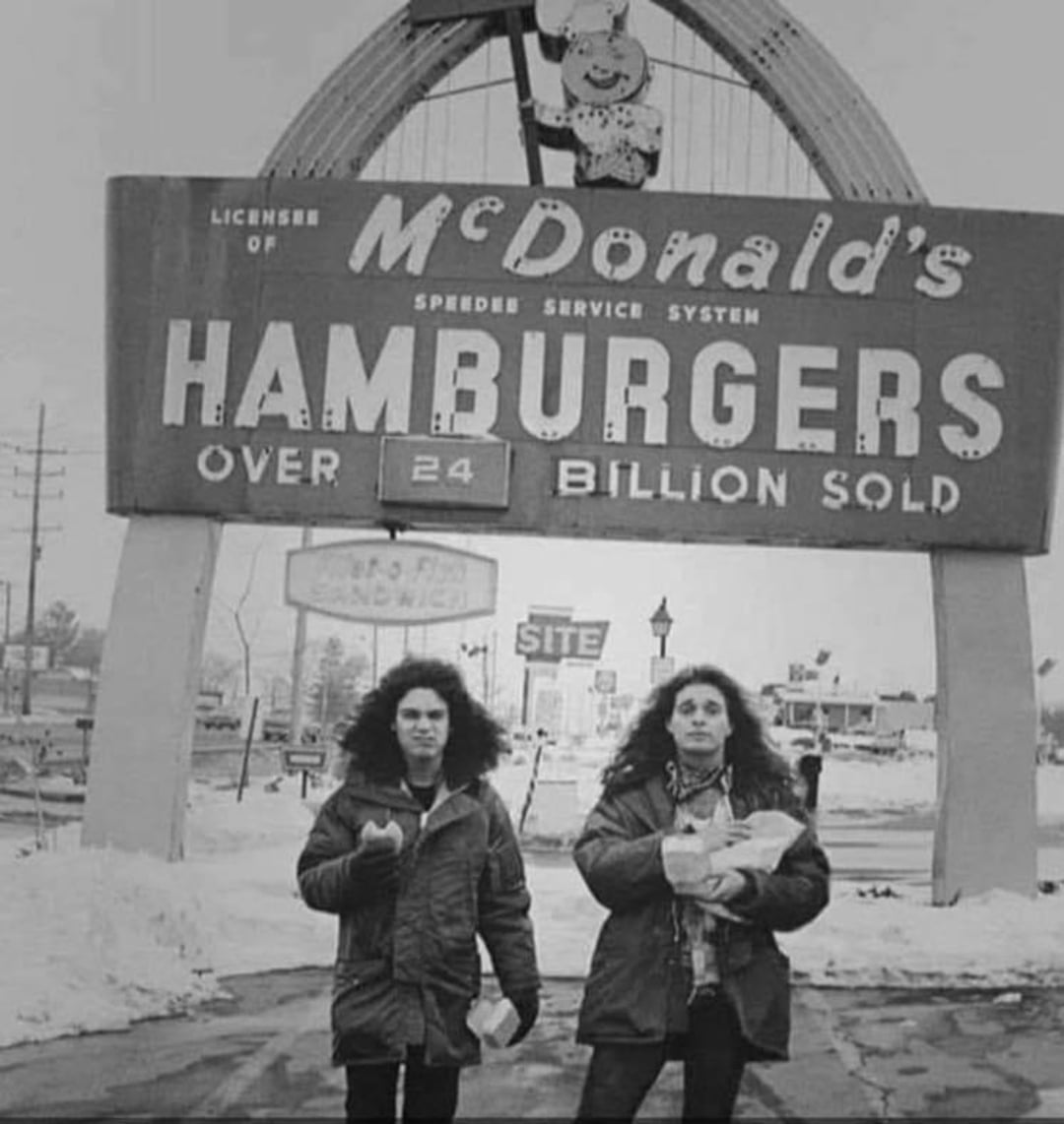 Two people with curly hair stand in front of a vintage McDonald's sign reading "HAMBURGERS Over 24 Billion Sold" on a snowy day, each holding food and drink, under the iconic golden arches.