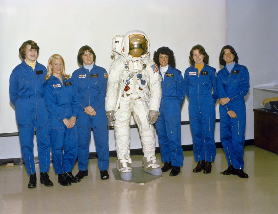 Six women in blue NASA jumpsuits stand next to a space suit display indoors, smiling for a group photo against a plain wall.