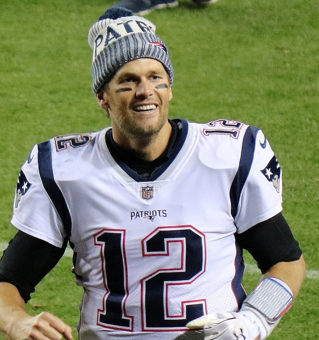 A football player in a white New England Patriots jersey with the number 12 and a blue-and-gray winter hat smiles while standing on a football field.