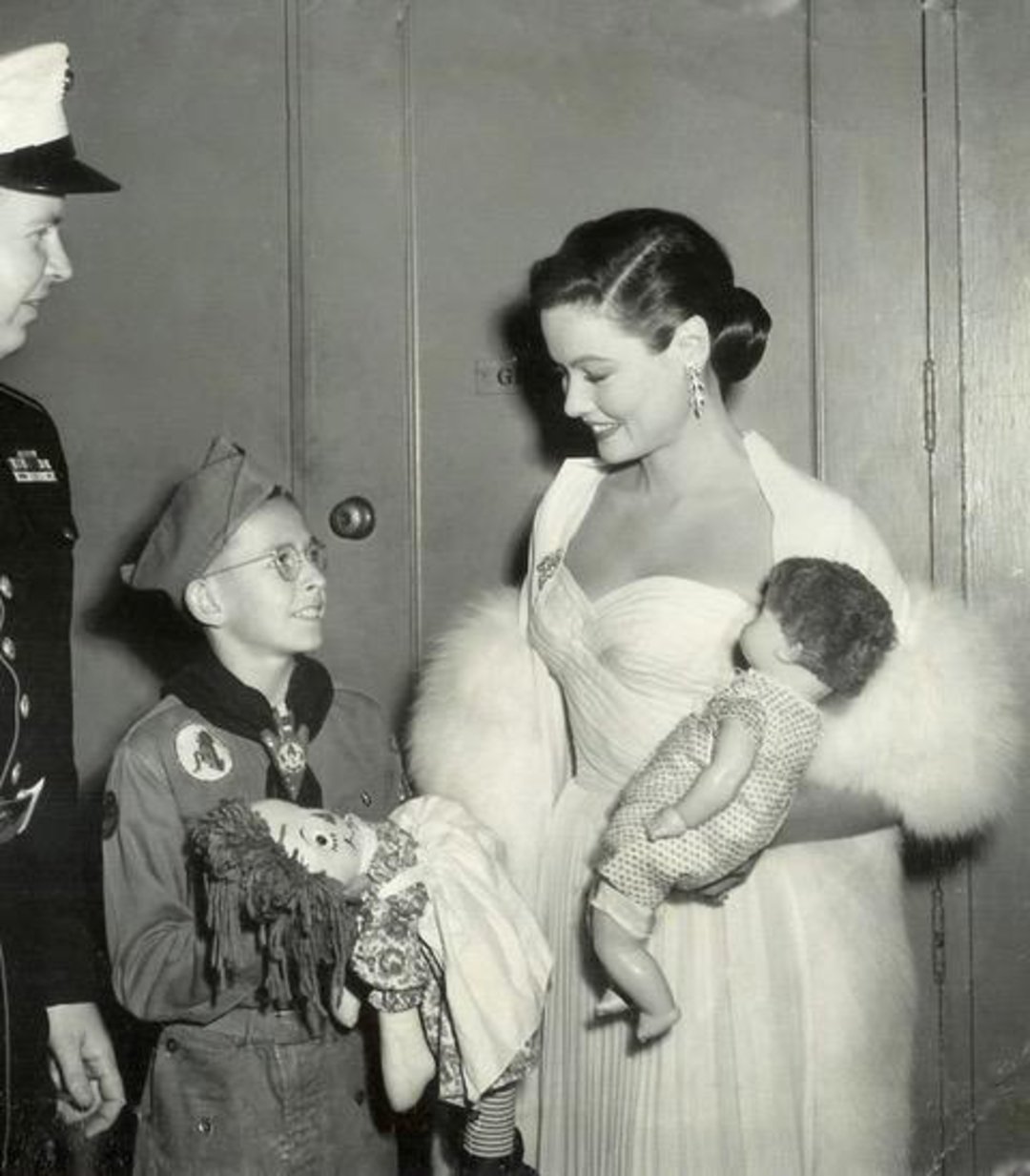 A woman in an elegant white gown holding a doll smiles at a young boy dressed as a scout, who is holding a rag doll. A man in a police uniform stands nearby. The group is gathered indoors, appearing cheerful.