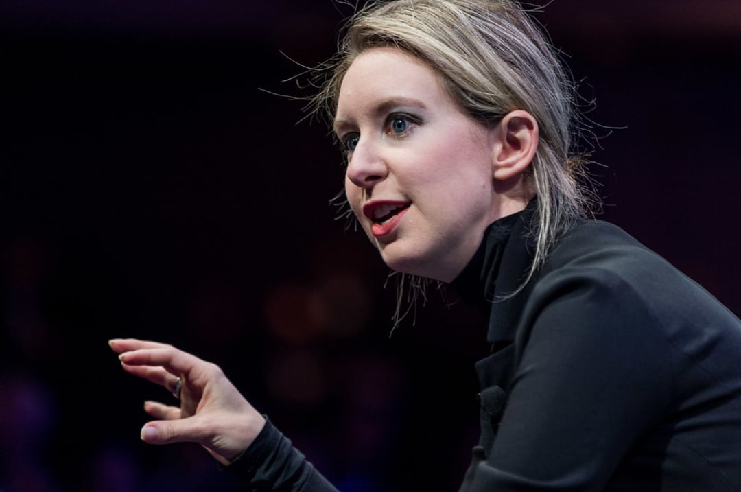 A woman with light skin and blonde hair pulled back is speaking animatedly, gesturing with her hand. She is wearing a black high-neck top and has a focused expression, with a dark, blurred background behind her.
