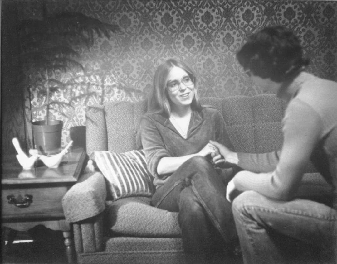 A black-and-white photo of two people sitting on a couch, shaking hands and smiling. One person has long hair and glasses, while the other has short hair. A side table with a plant and decorative items is nearby.