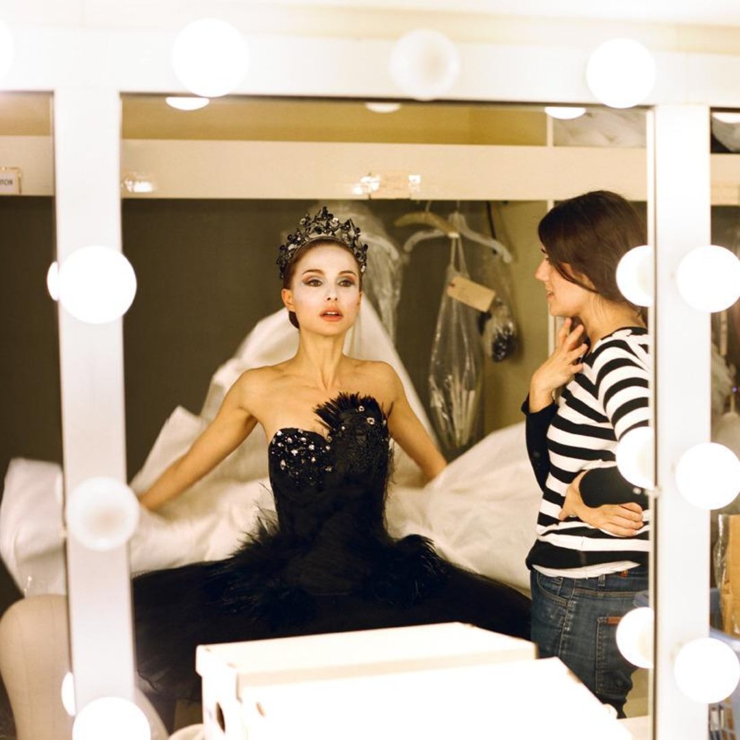 A woman in a black, feathered ballerina costume and jeweled crown stands with arms slightly raised in front of a mirror lit by round bulbs, while another woman in a striped shirt looks on.