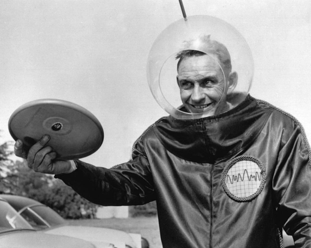 A man in a shiny jacket with a futuristic emblem and a clear, round helmet holds up a frisbee, smiling. The black-and-white photo has a retro, science fiction vibe.