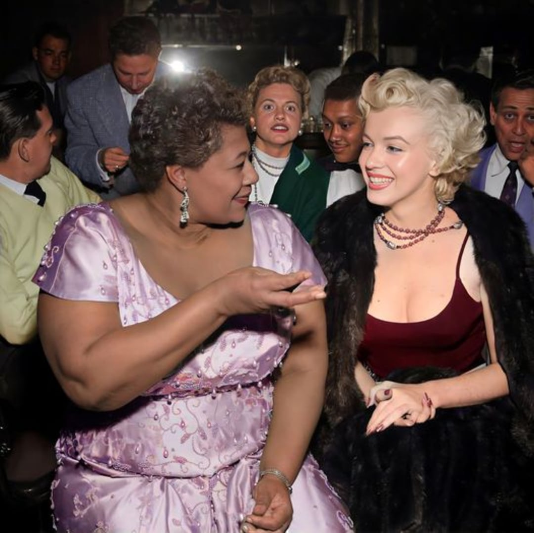 Two women, one wearing a lavender dress and the other a black fur shawl over a red dress, sit closely together, smiling and talking in a crowded, elegant setting with several people in the background.