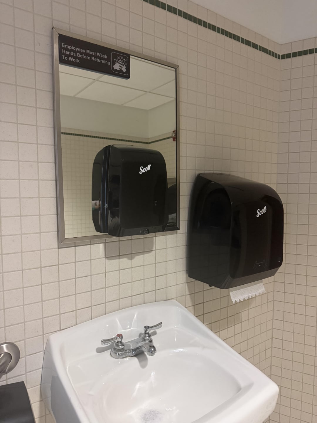 A restroom sink with a silver faucet, a wall mirror, a paper towel dispenser, and a soap dispenser. A sign above the mirror reads, “Employees must wash hands before returning to work.”