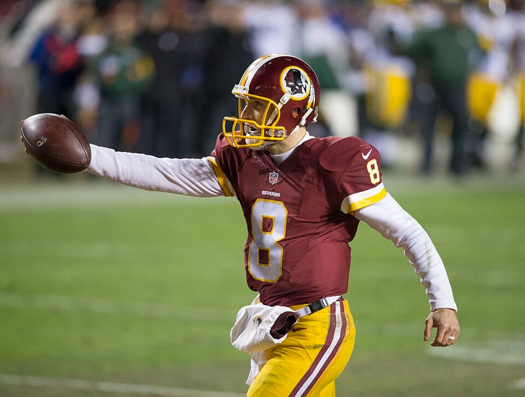 A football player in a maroon and gold uniform with the number 8 and a feathered head logo extends his arm holding a football during a game on a lit field. The stands and other players are visible in the background.
