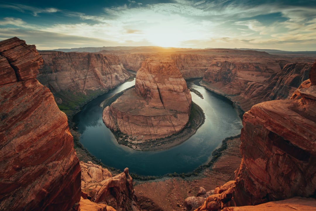 A panoramic view of Horseshoe Bend at sunset, with the Colorado River curving around a massive rock formation, surrounded by dramatic red cliffs under a partly cloudy sky.