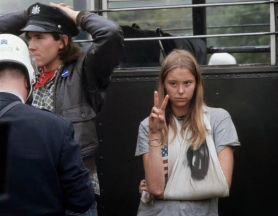 A young woman with a bandaged arm in a sling gives a peace sign, standing beside a man in a leather jacket and police officers near a bus, likely during a protest or demonstration.