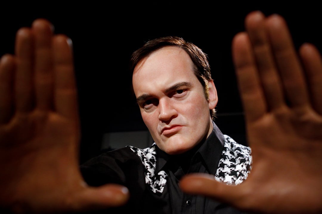 A man with short dark hair and a serious expression holds his hands up in front of him, forming a rectangular frame with his fingers, as if visualizing a scene. He wears a black shirt with a patterned jacket.